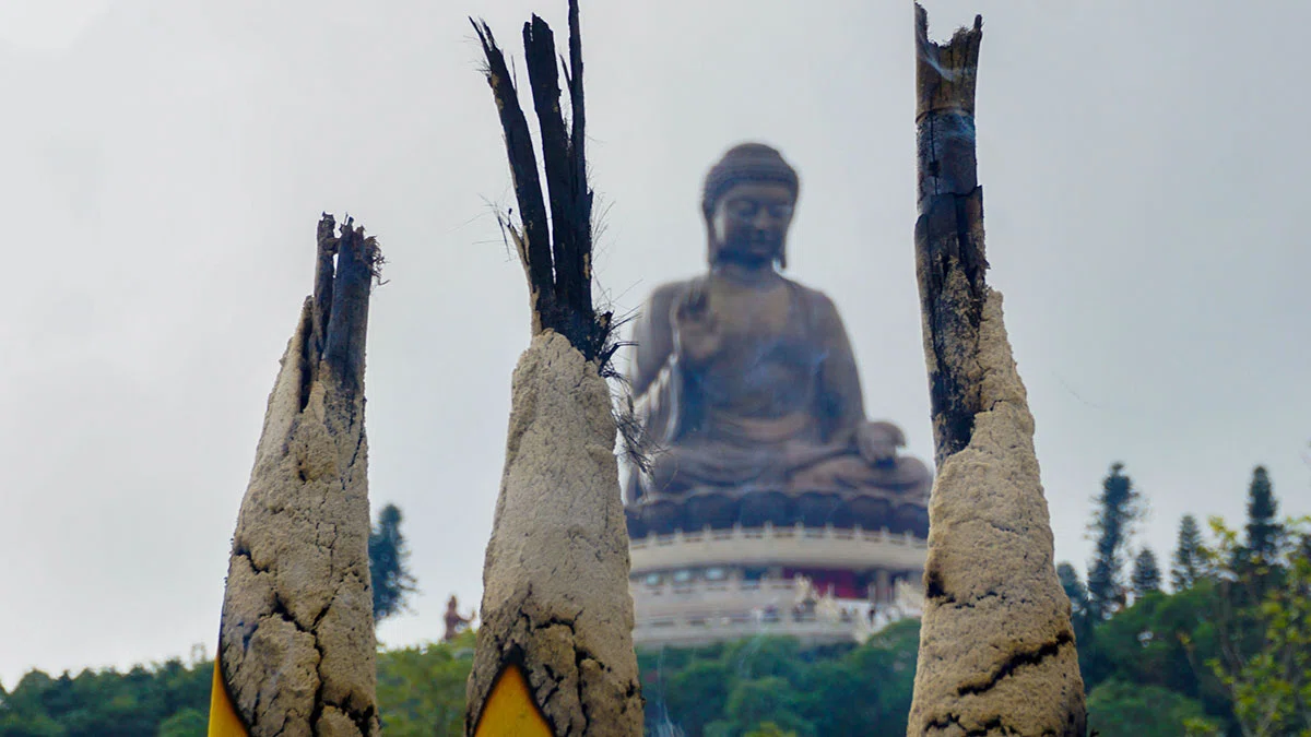 Incense sticks burn in foreground with large Buddha statue in background.