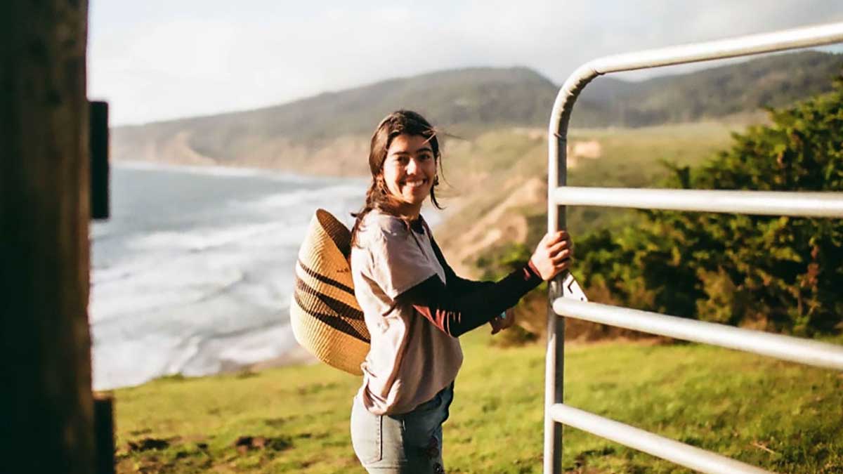 student smiles for camera holding a gate open on a  cliffside by the ocean